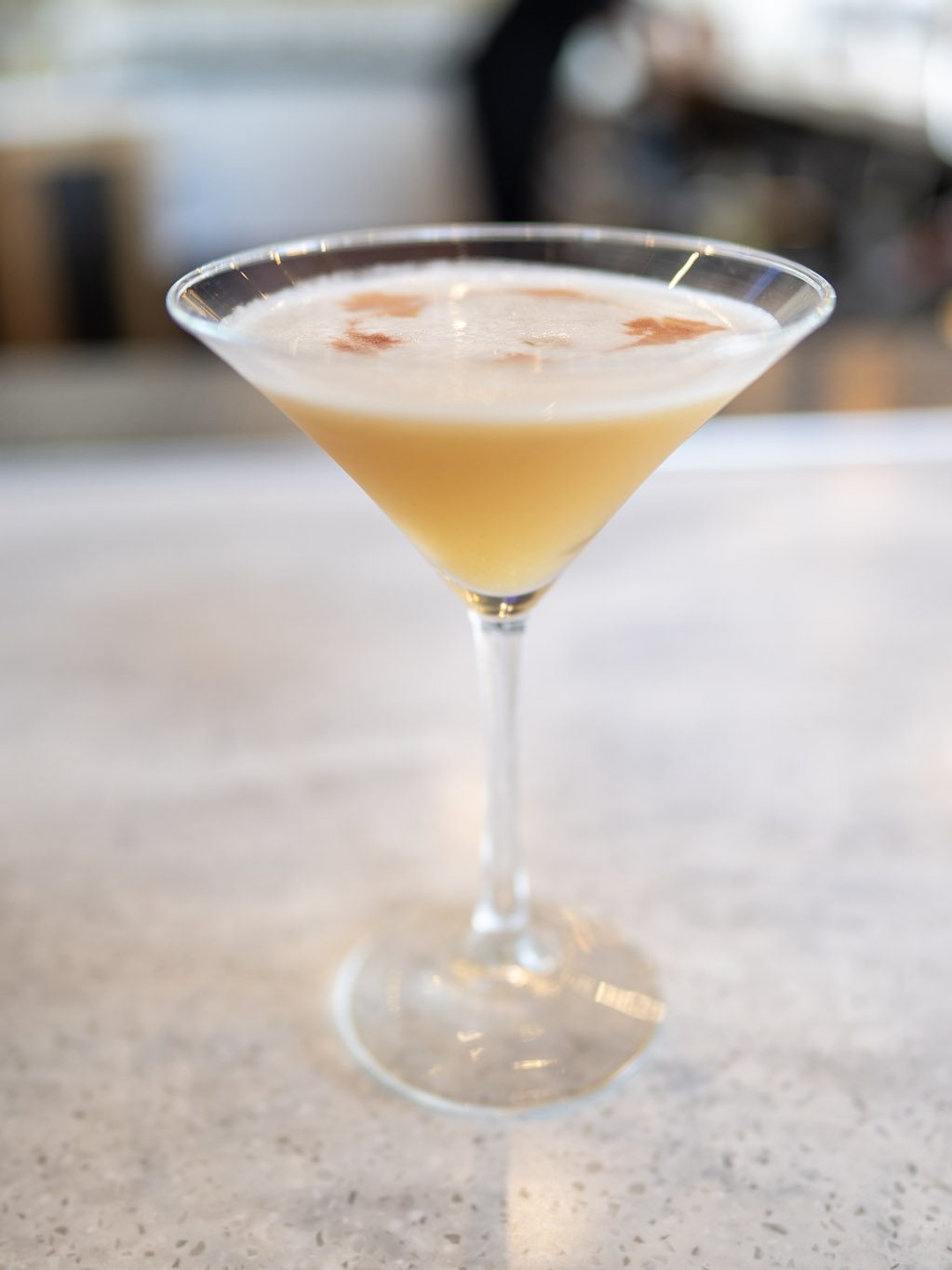 A pale, frothy cocktail served up in a martini glass and dusted with nutmeg, on the marble bar with the back bar softly out of focus