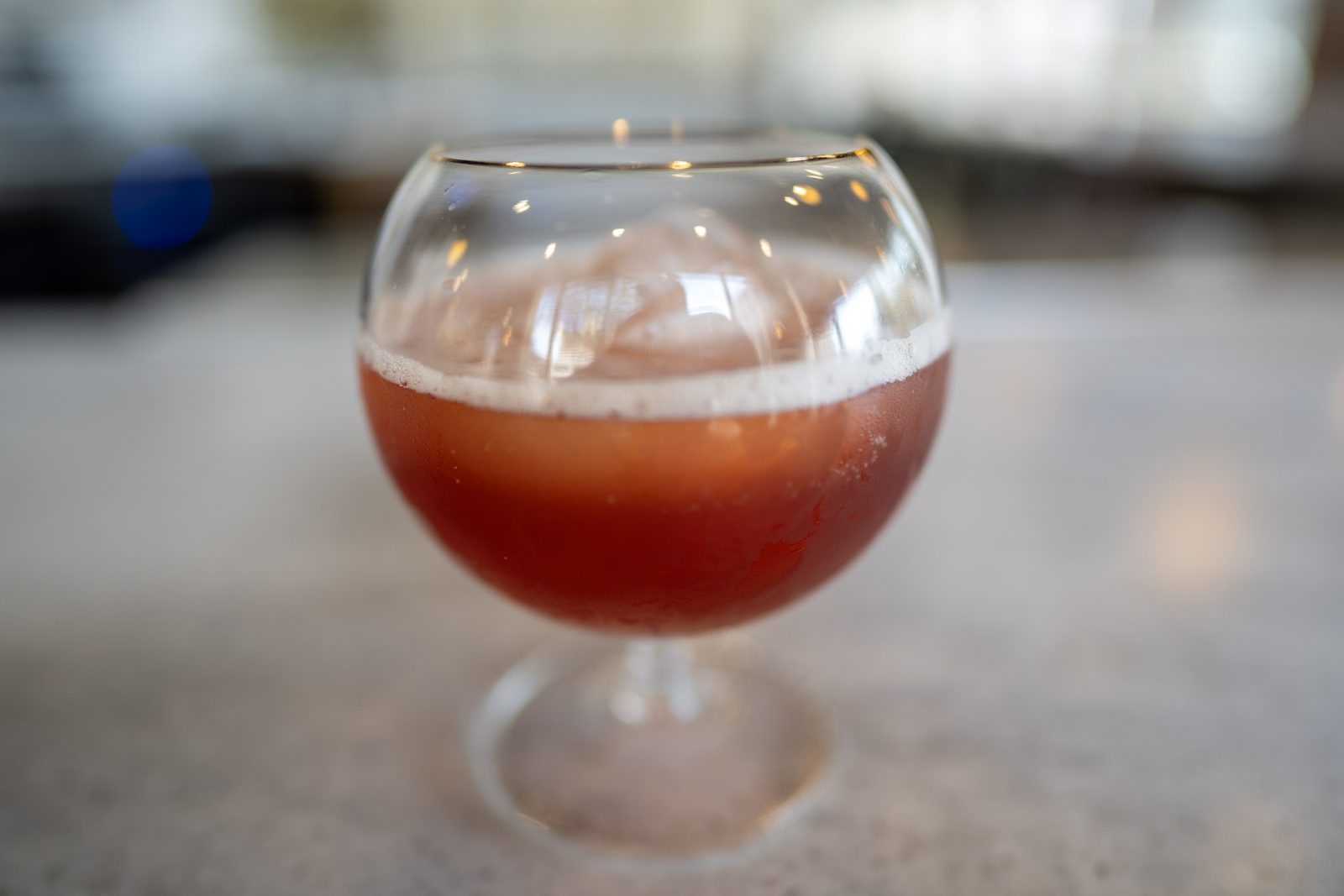A deep red cocktail over ice in a snifter on the marble bar, the back bar softly out of focus