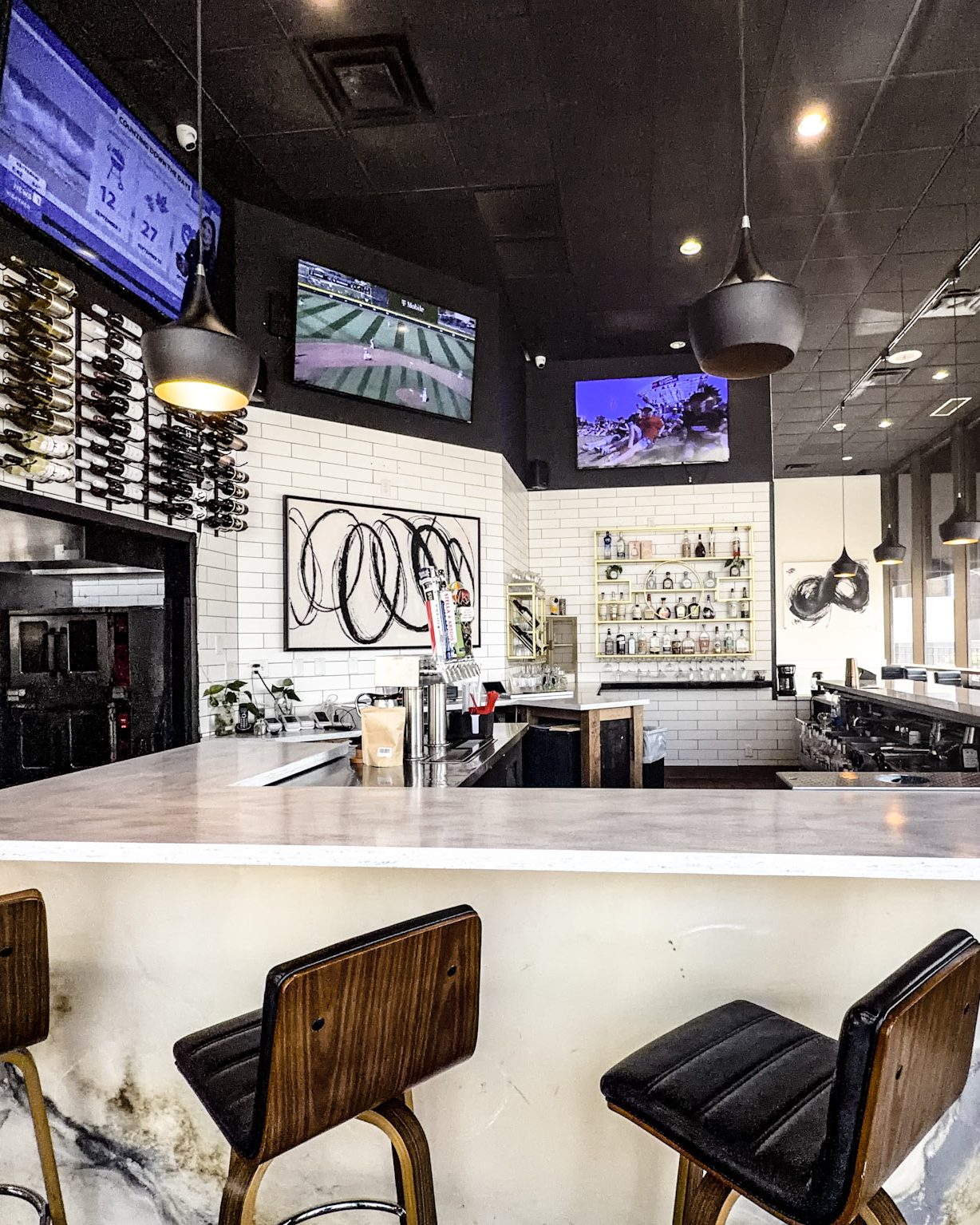 The Flavor House bar in daylight: a marble-topped counter with leather stools, black pendant lights, a wall-mounted wine rack, and the back bar shelves against white subway tile