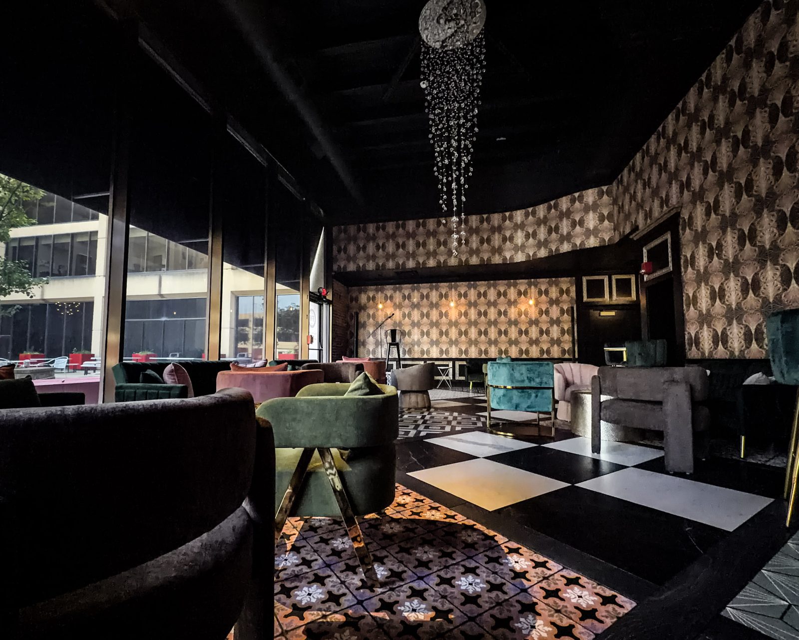 The Flavor House lounge: velvet armchairs in green, blush, and teal on a black-and-white checkerboard floor, patterned wallpaper, and a crystal chandelier overhead